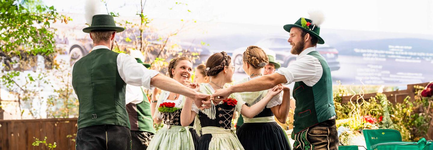 Airbräuwiesn A group of dancers in Bavarian traditional dress holding hands in a circle and dancing cheerfully. The scene is lively and traditional, reflecting Bavarian culture and the festive atmosphere at Munich Airport.