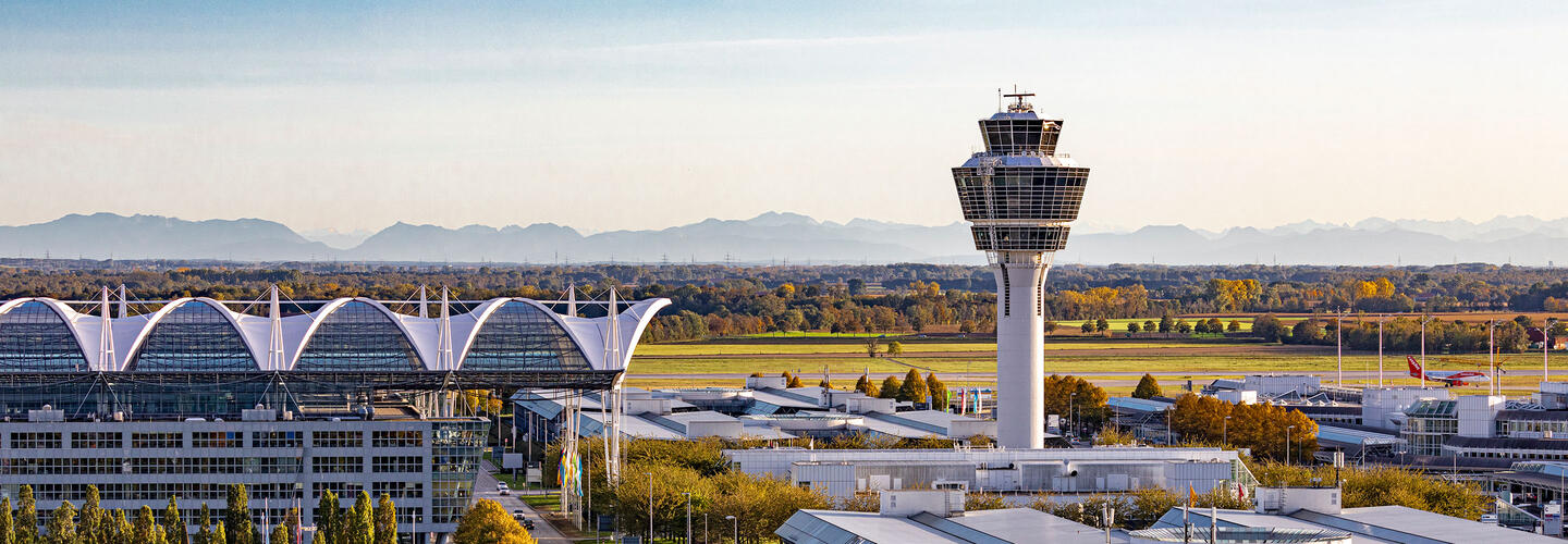Airport tower in the warm evening light The tower of Munich Airport stands in the golden evening light against an autumnal landscape and the mountains in the background. The atmosphere is calm, warm, and inviting.
