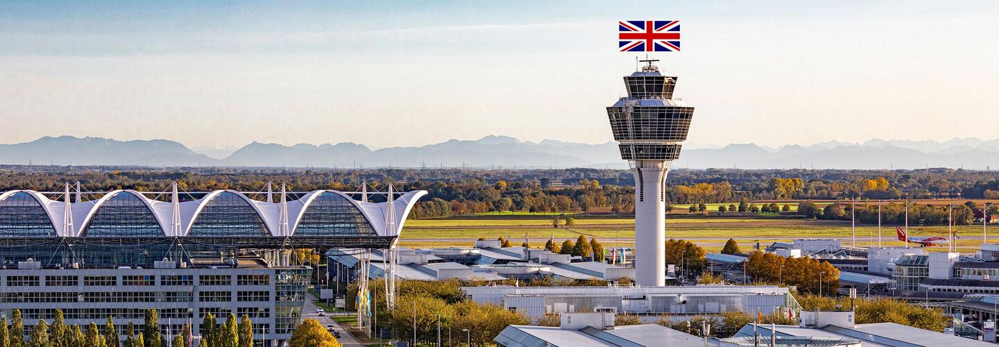 Trainings und Schulungen am Flughafen München Der Tower des Flughafens München ist in den Farben des Union Jack beleuchtet. Vor ihm liegen die ikonischen Terminaldächer und die herbstliche Landschaft, im Hintergrund ein sanftes Alpenpanorama. Das Bild wirkt feierlich und international.