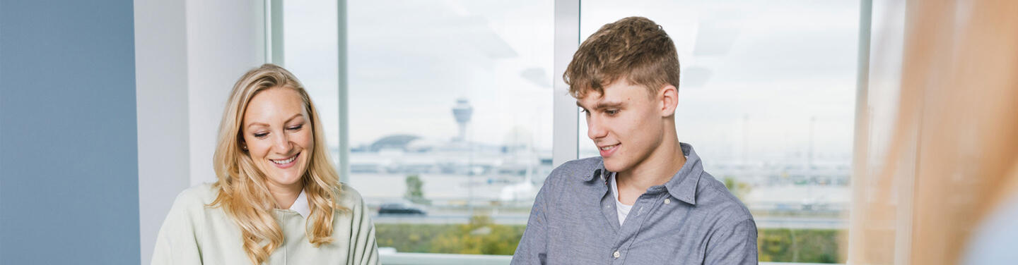 Bachelor Wirtschaftsinformatik am Flughafen München Eine junge Frau und ein junger Mann sitzen freundlich lächelnd im Gespräch an einem Arbeitsplatz mit Fensterblick auf den Flughafen München; ruhige, kooperative Lernatmosphäre.