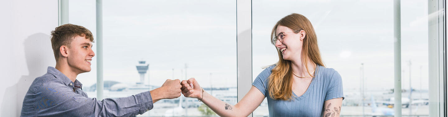 Fachinformatiker Systemintegration Zwei junge Personen geben sich lächelnd einen Fistbump vor einem Fenster mit Blick auf den Flughafen München; freundliche, offene Lern- und Teamatmosphäre.
