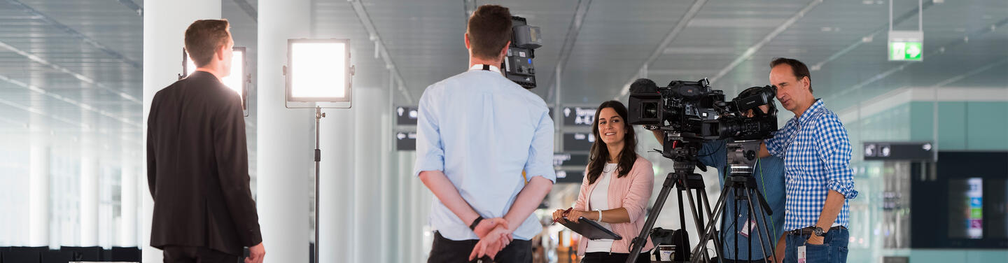 Munich Airport – Media Filming Setup Media crew inside Munich Airport with two video cameras on tripods, lighting setup and an interview subject, in the terminal area.