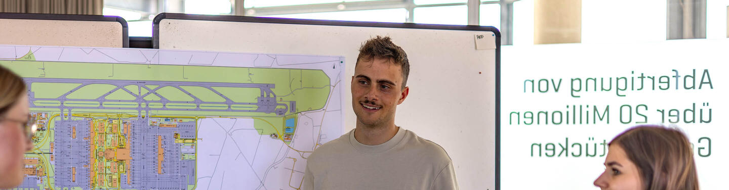 Ausbildung Immobilienkaufmann/-frau am Flughafen München Ein Auszubildender steht vor einer großen Planzeichnung mit Geländeübersicht des Flughafens München und spricht mit Kolleginnen; professionelle, konzentrierte und kooperative Arbeitsatmosphäre.