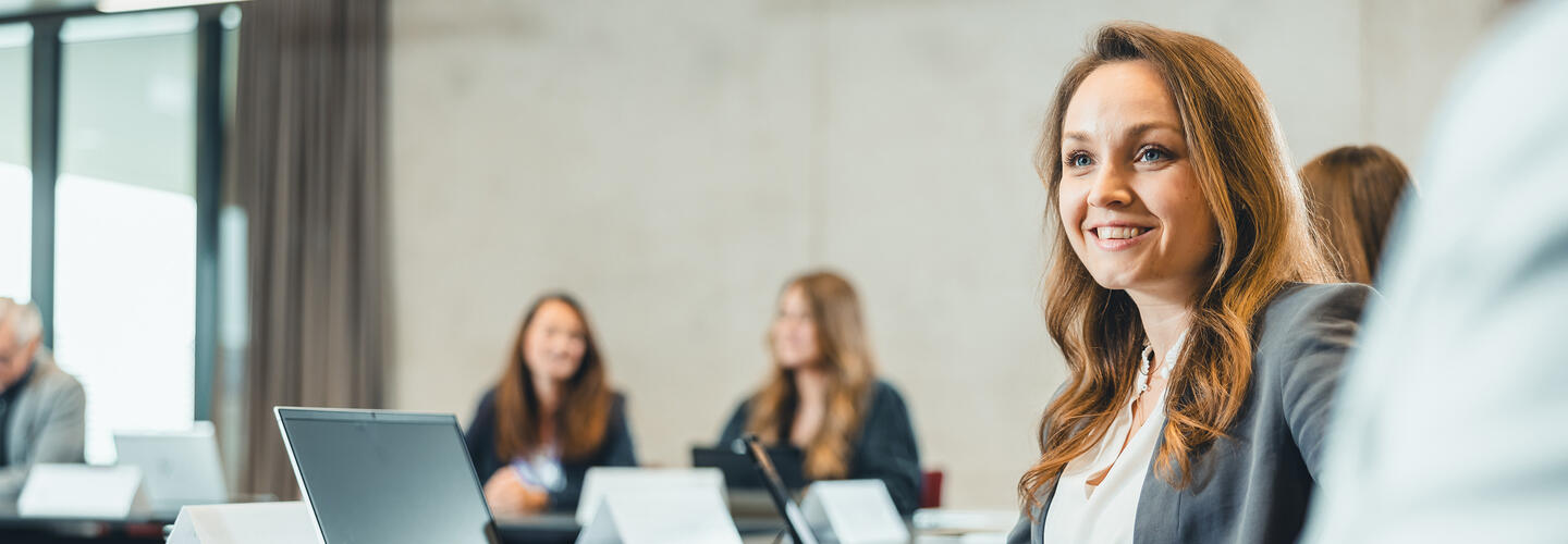 AirportAcademy des Flughafens München In einem hellen, modernen Besprechungsraum sitzt eine junge Frau lächelnd an ihrem Laptop, während Kolleginnen und Kollegen im Hintergrund konzentriert zuhören. Die Stimmung wirkt professionell, offen und freundlich.