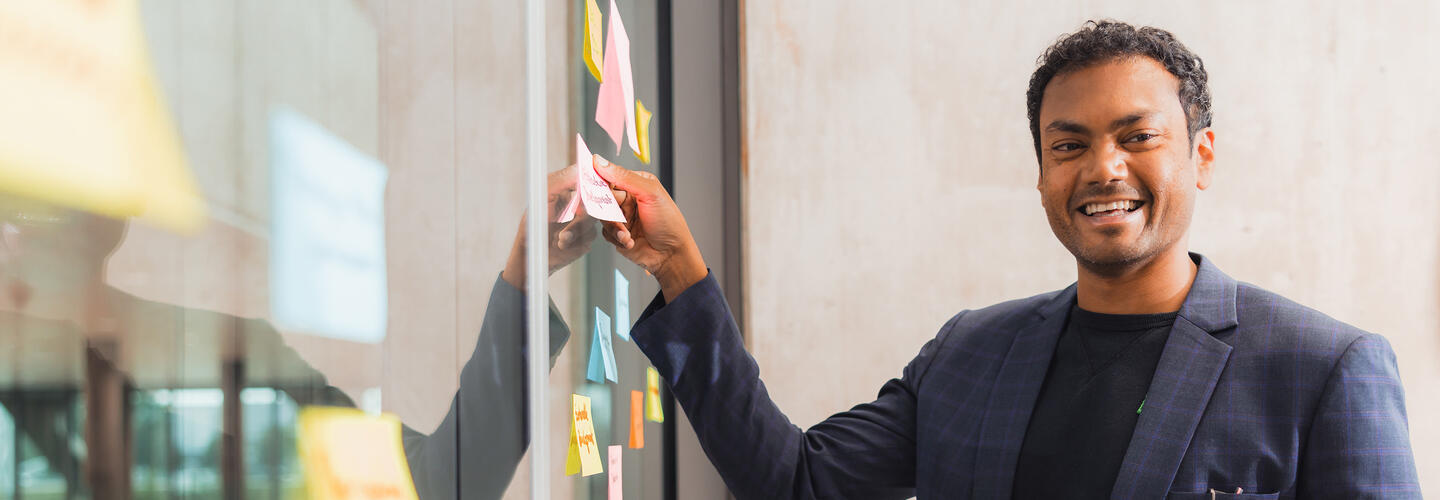 Trainings at Munich AirportAcademy An employee stands in front of a glass wall covered with colorful Post-it notes and points to an idea with a pen while smiling. The scene conveys dynamism, creativity, and teamwork in a modern work environment.