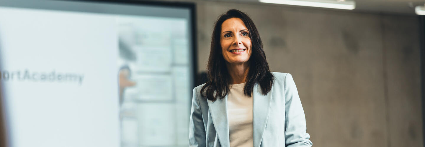 Die AirportAcademy am LabCampus Eine Trainerin steht selbstbewusst vor einer Präsentationsleinwand und spricht zu einer Gruppe. Die Umgebung ist hell, professionell und vermittelt den Eindruck einer Schulung oder Weiterbildungssituation.