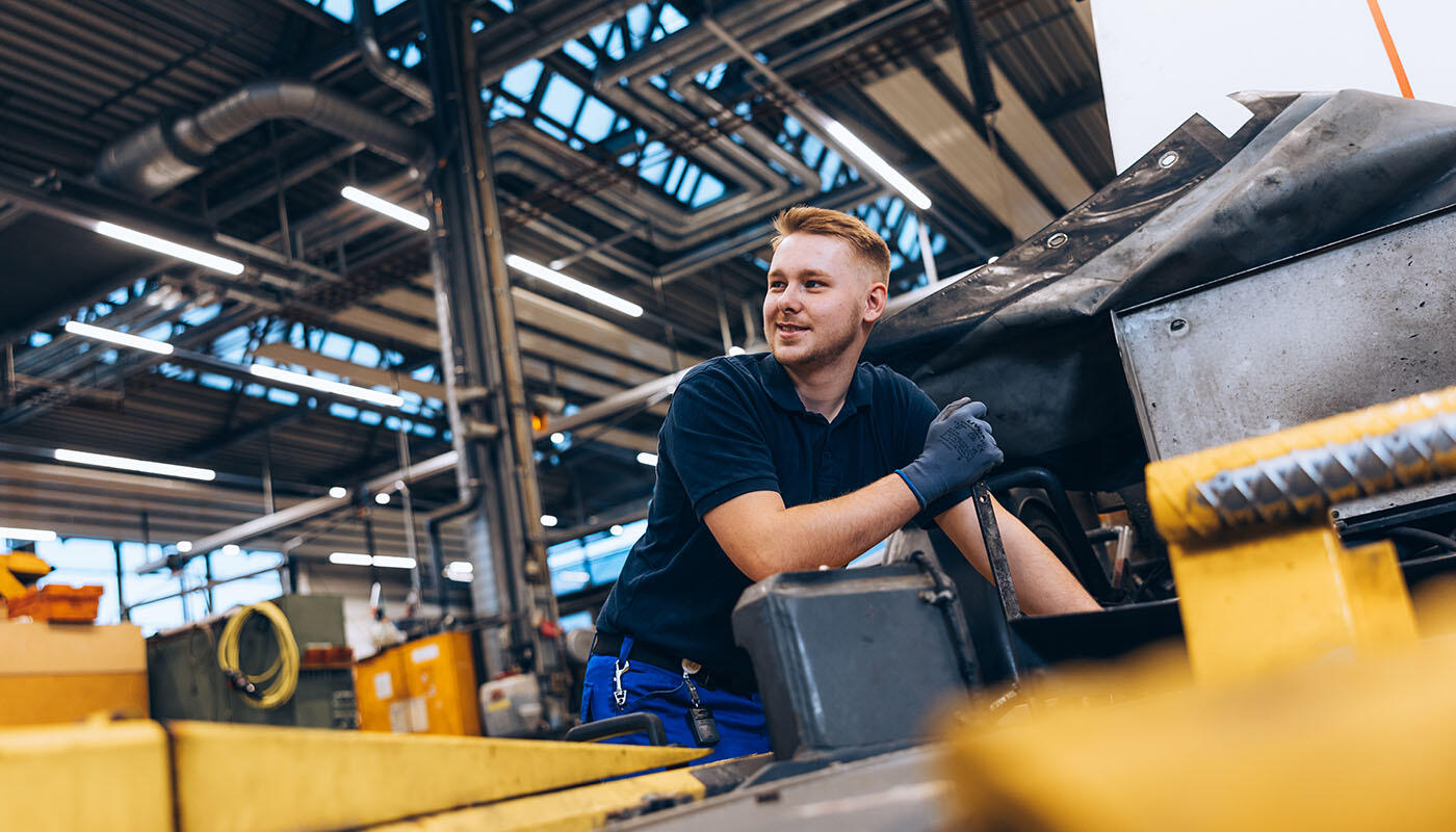 Mechatroniker in der Kfz-Werkstatt vom Fahrzeugmanagement am Flughafen München Person arbeitet in einer Kfz-Werkstatt an einer großen Maschine, umgeben von industriellen Werkzeugen.