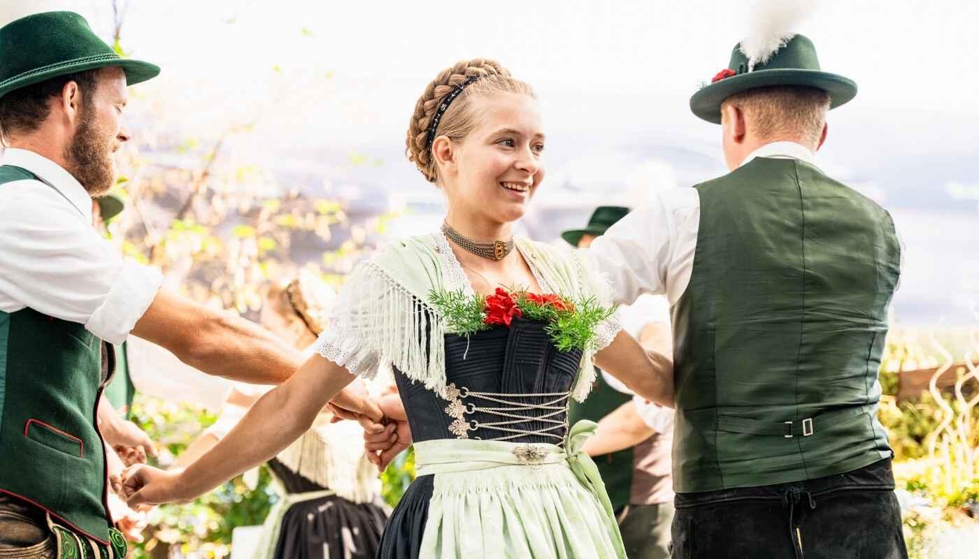 Bavarian traditional dress and folk dance A young woman in a dirndl smiles brightly as she dances in a circle with others in traditional attire, hands joined, sunlight and joyful tradition in the air.