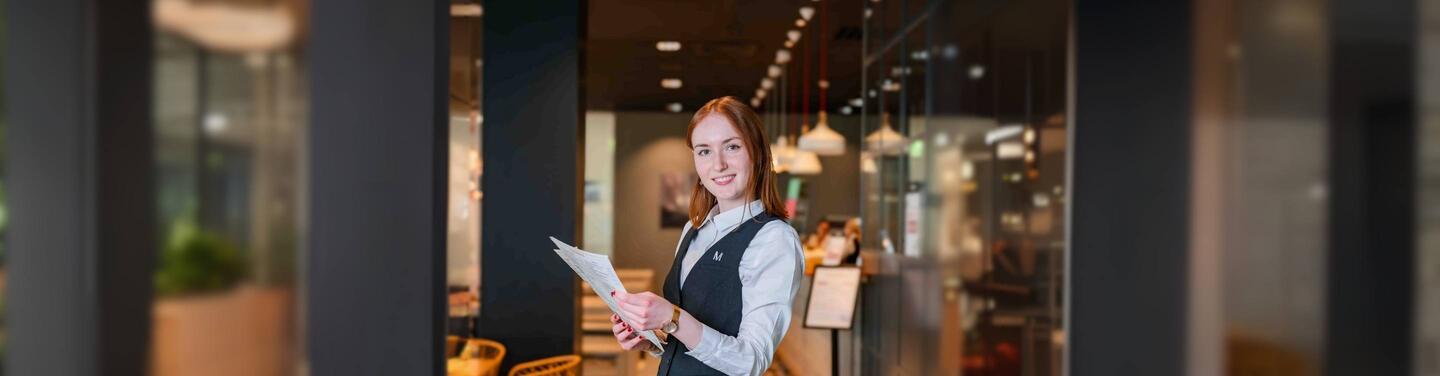 Karriere bei Allresto – Service-Team Junge Service-Mitarbeiterin hält eine Speisekarte und lächelt, im Hintergrund ein modern beleuchteter Restaurantbereich.