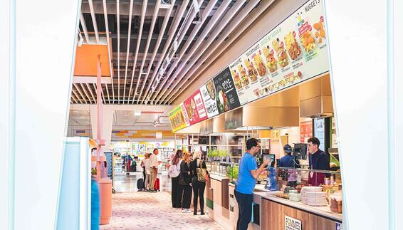 Bestelltheke des Restaurants 4 FOOD mit verschiedenen Stationen - Pommesfreunde, Asia, Nudeln und Pizza. Einige Passagiere zum Teinl mit kleinen Koffern stehen vor der Theke, hinter der Theke befinden sich zwei Mitarbeiter.