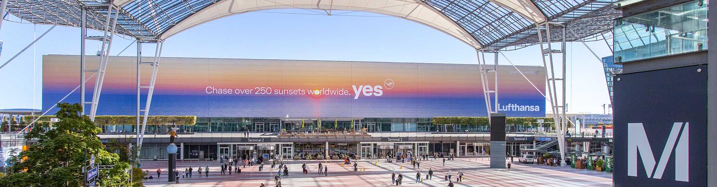 Lufthansa West Facade - Sunset Die Westfassade des Terminals 2 am Flughafen München ist mit einem Motiv der Lufthansa bespielt. Darauf ist ein Sonnenaufgang zu sehen sowie der Schriftzug "Chase over 250 sunsets worldwide - yes".