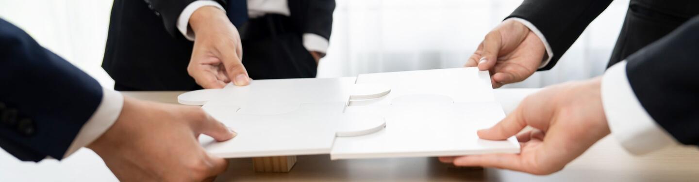 Vergabe- und Beschaffungsportal Hände von vier verschiedenen Personen fügen große weiße Puzzleteile zusammen, als Keyvisual für Teamarbeit und Vernetzung im Vergabeportal