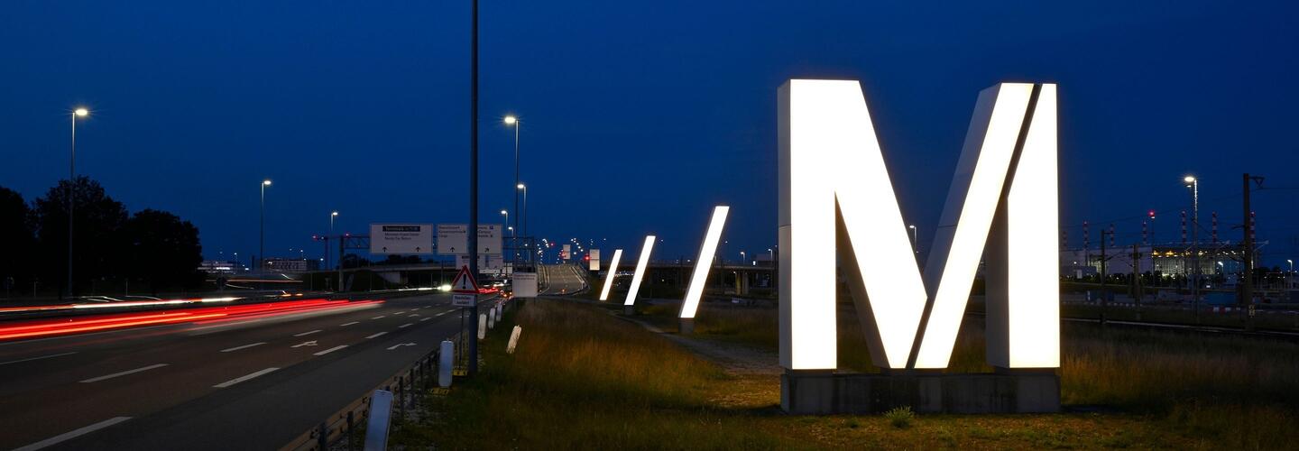 Evening atmosphere at Munich Airport - Access to the airport The iconic, glowing white /M sculpture stands in the foreground, with car lights stretching out behind it in long lines through the twilight. The motif conveys the modernity and recognizability of Munich Airport.