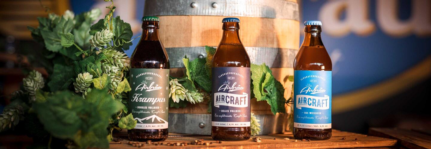 Airbräu Bierflaschen auf Holzfass Drei unterschiedlich etikettierte Airbräu-Bierflaschen stehen dekorativ auf einem Holzfass, umgeben von frischem Hopfen und grünen Blättern. Warmes, einladendes Licht erzeugt eine gemütliche Brauerei-Atmosphäre.