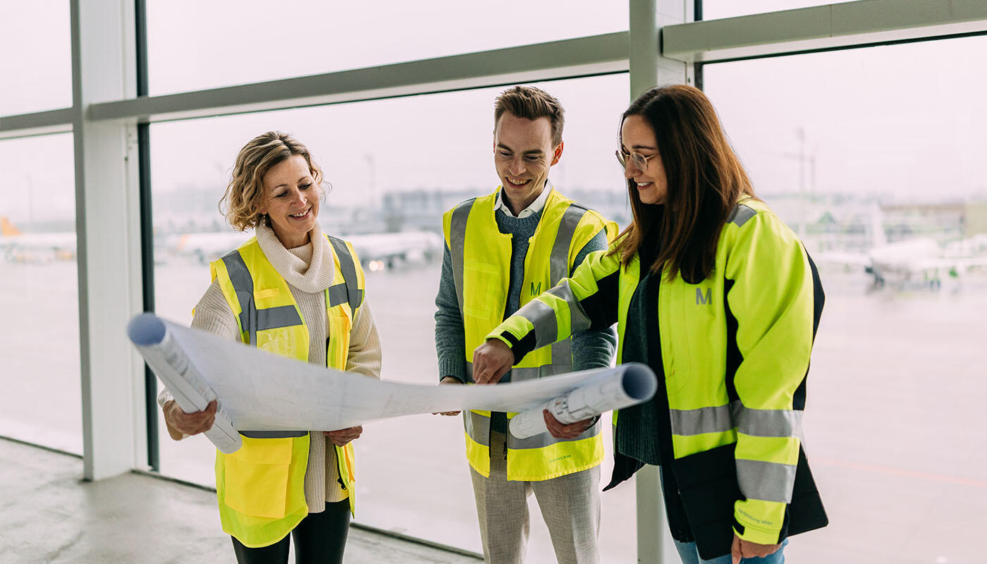 Bauingenieur Christian Einblick in die Jobwelt eines Bauingenieurs am Flughafen München: Drei Mitarbeitende in Warnwesten betrachten gemeinsam Baupläne mit Blick auf das Flughafenvorfeld.