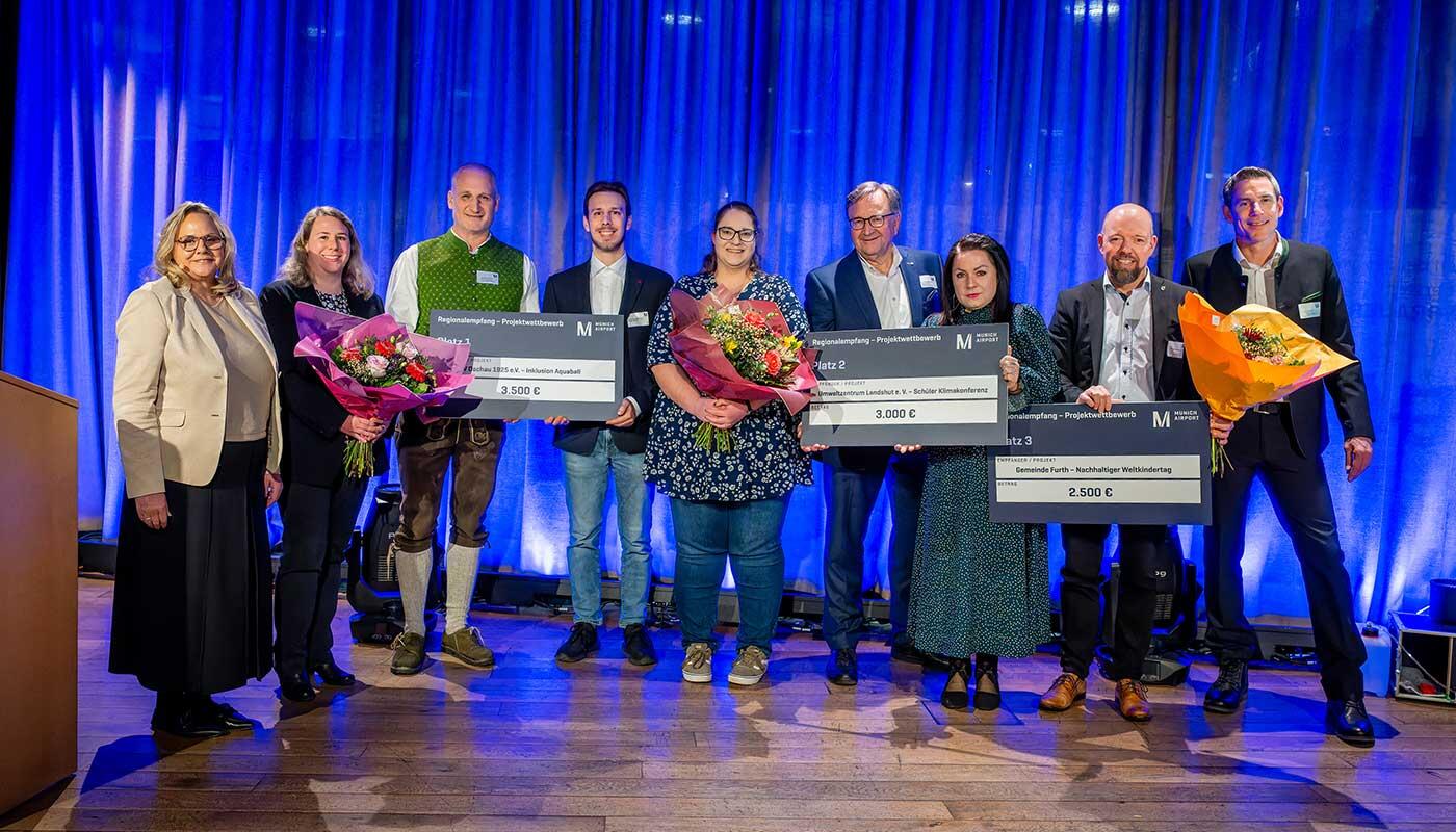 Flughafen München ehrt Gewinner des Projektwettbewerbs Gruppenfoto beim Regionalempfang 2025: Mehrere Personen stehen auf einer Bühne vor blau beleuchtetem Hintergrund, halten große symbolische Spendenschecks und Blumensträuße und werden für ihr ehrenamtliches Engagement in der Region von der Flughafen München GmbH ausgezeichnet.
