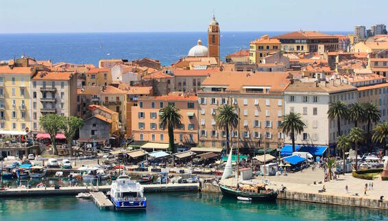 Blick auf den Hafen von Ajaccio auf Korsika mit Segelbooten, Palmen und pastellfarbenen Häusern der Altstadt. Im Hintergrund liegt das türkisfarbene Mittelmeer und die mediterrane Küste.