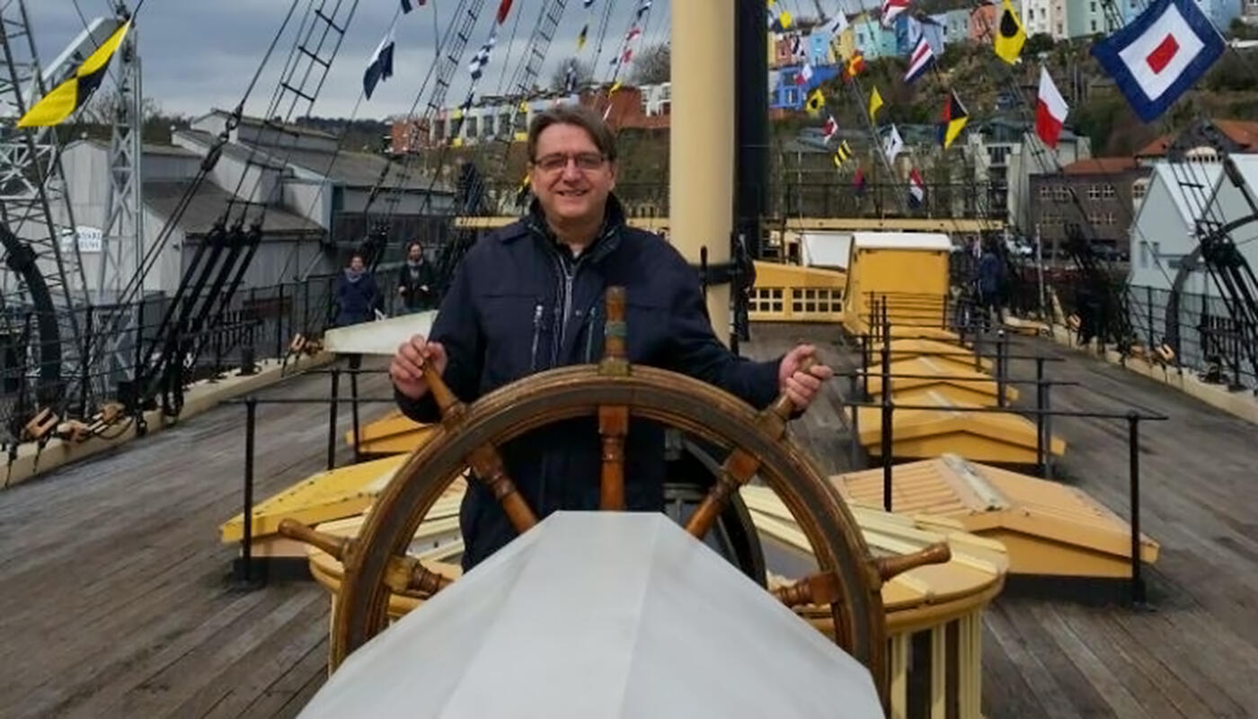 Insider Ingo Ansbach Ingo Ansbach steht lachend am großen hölzernen Steuerrad auf dem Deck der SS Great Britain in Bristol; bunte Signalflaggen hängen an den Masten, im Hintergrund sind Besucher, Hafenanlagen und farbige Häuser am Hang zu sehen.