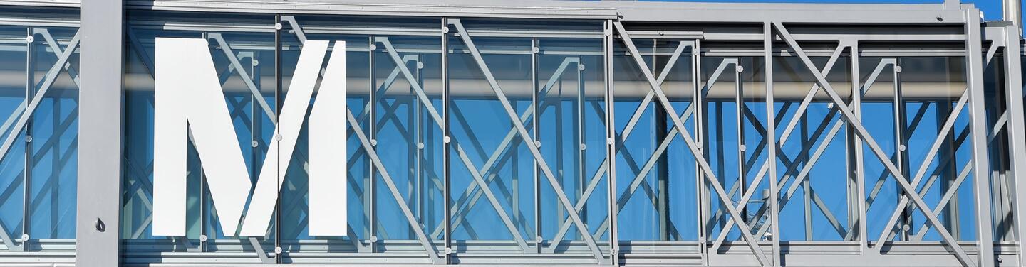 Munich Airport Large white letter “M” mounted on a glass façade with visible metal framework; a blue sky is visible in the background, and the scene appears modern and clear.