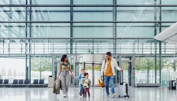 Mother and father with two young boys standing with travel luggage inside a modern airport building with glass facade in the background