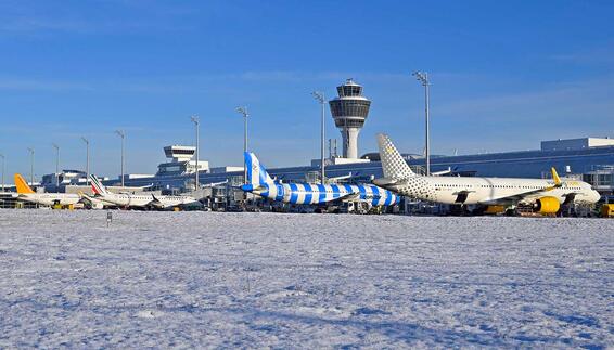 Verschneites Vorfeld am Flughafen München mit mehreren geparkten Passagierflugzeugen und dem Tower im Hintergrund an einem klaren Wintertag.