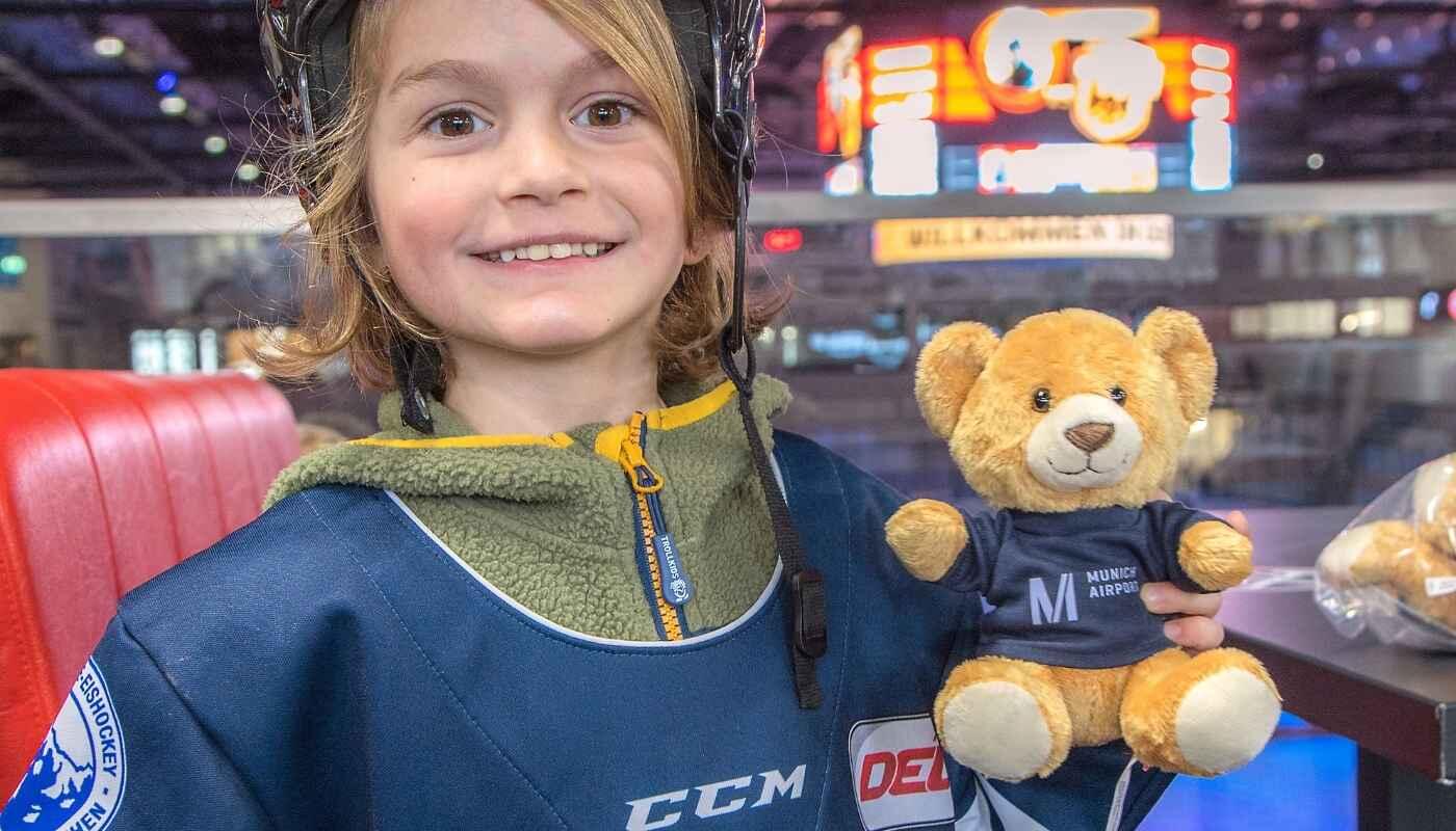 Kind beim Benefiz-Eishockeyspiel mit Teddy – Teddy Bear Toss für den guten Zweck in Landshut Lächelndes Kind in Eishockeyausrüstung sitzt in einer Arena und hält einen Teddybären während eines Benefiz-Eishockeyspiels mit Teddy-Bear-Toss Aktion.