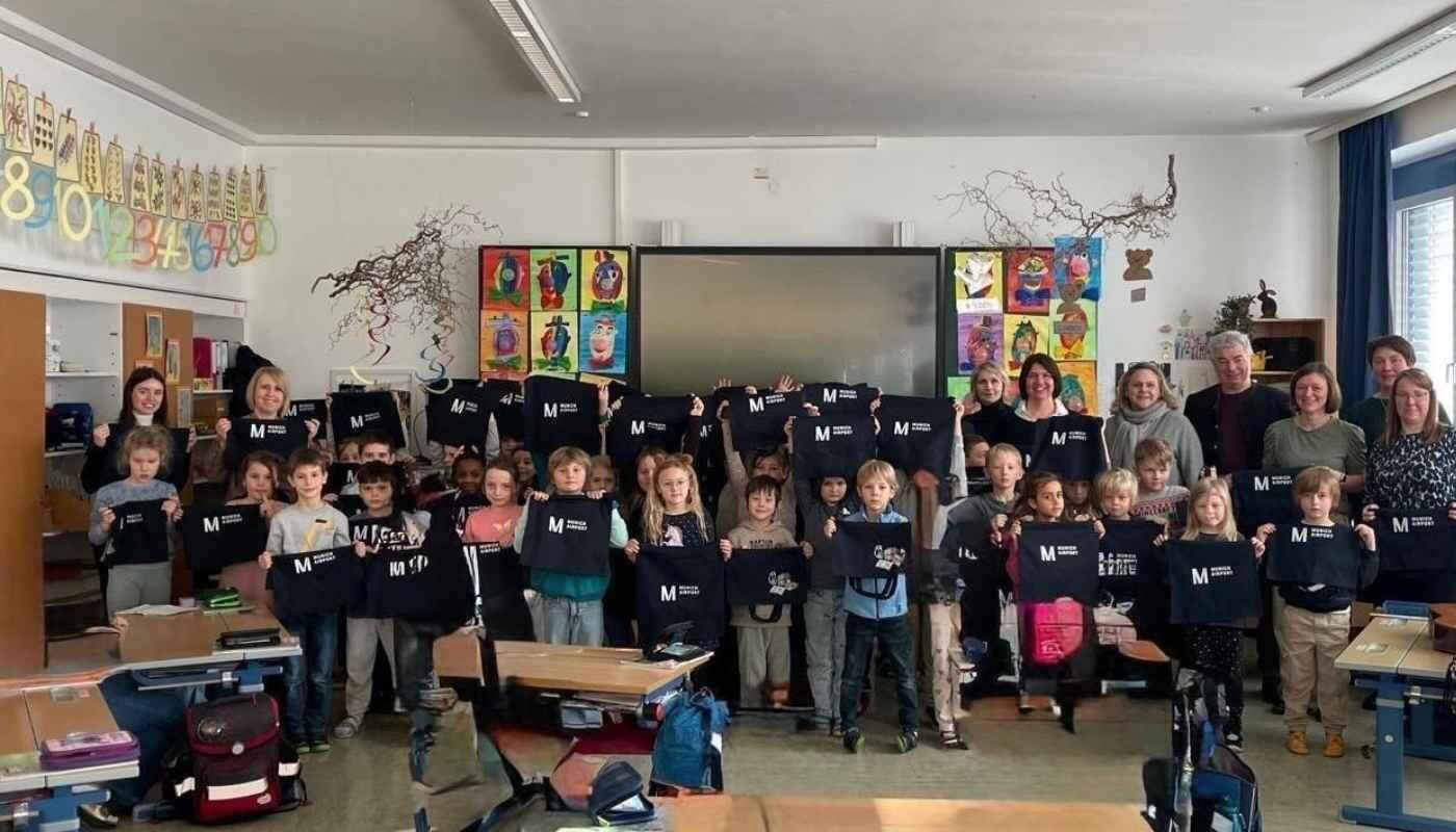 Büchertaschen vom Flughafen München für Erstklässler – Leseförderprojekt der Grundschule Velden Eine erste Klasse der Grundschule Velden steht gemeinsam in einem Klassenzimmer und hält gespendete Büchertaschen in die Kamera. Hinter den Kindern stehen Lehrkräfte und erwachsene Unterstützer. Schulbänke, Schulranzen und farbenfrohe Lernposter sind sichtbar; das Bild zeigt eine offizielle Übergabeaktion im Rahmen einer Leseförderung durch den Flughafen München.