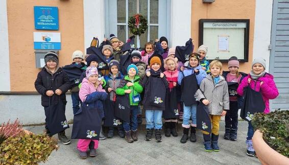 Erstklässler der Grundschule Velden stehen vor dem Schulgebäude und halten vom Flughafen München gespendete dunkle Büchertaschen in die Kamera; die Kinder tragen Winterkleidung und lächeln bei einer offiziellen Übergabeaktion im Außenbereich der Schule.