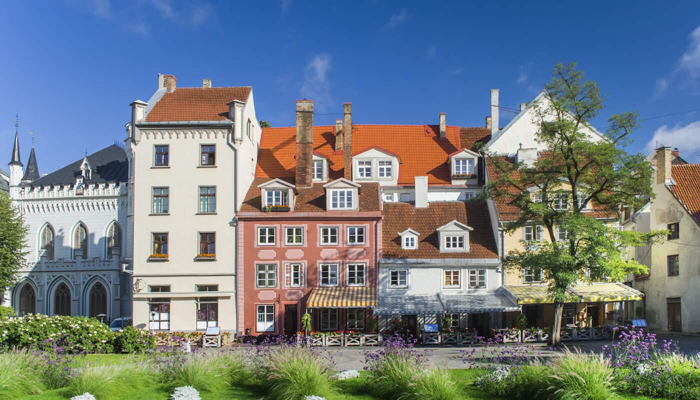 Altstadt von Riga, Lettland Farbenfrohe, historische Häuser mit roten Ziegeldächern in der Altstadt von Riga, Lettland. Im Vordergrund liegt eine gepflegte Grünanlage mit Blumenbeeten und violetten Blüten. Der Himmel ist klar und blau.