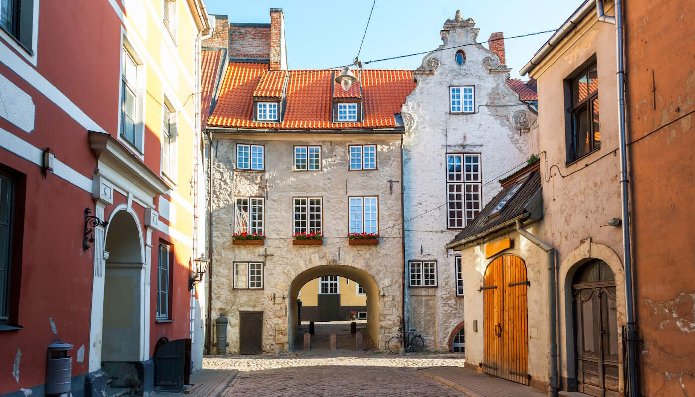 Kleine Gasse in Riga, Lettland Schmale Gasse mit Kopfsteinpflaster in Riga, Lettland. Beidseitig stehen alte, teils pastellfarbene Häuser mit kleinen Fenstern und Holztüren. In der Mitte des Hauses am Ende der Gasse befindet sich ein Torbogen, der den Blick in einen dahinterliegenden Bereich freigibt. Der Himmel ist klar und blau.