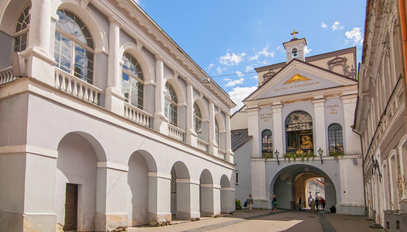 Tor der Morgenröte in Vilnius, Litauen Tor der Morgenröte in Vilnius, Litauen, mit weiß verputzten Gebäuden und Arkadengängen. In der Mitte ist ein Tor mit einer kleinen Kapelle darüber, geschmückt mit religiösen Symbolen und goldenen Details. Menschen gehen durch den Torbogen, darüber spannt sich ein blauer Himmel mit wenigen Wolken.