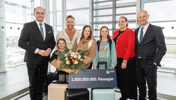 A group with suitcases at the airport is holding a sign for the billionth passenger and a bouquet of flowers