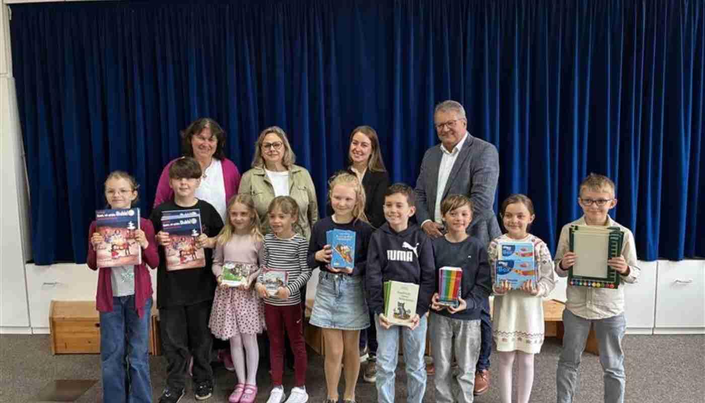 Grundschule Bockhorn Grundschulkinder halten ihre neuen Bücher in den Händen und blicken mit vier Erwachsenen gemeinsam vor einer Bühne lächelnd in die Kamera. Im Hintergrund ein dunkelblauer, geschlossener Bühnenvorhang
