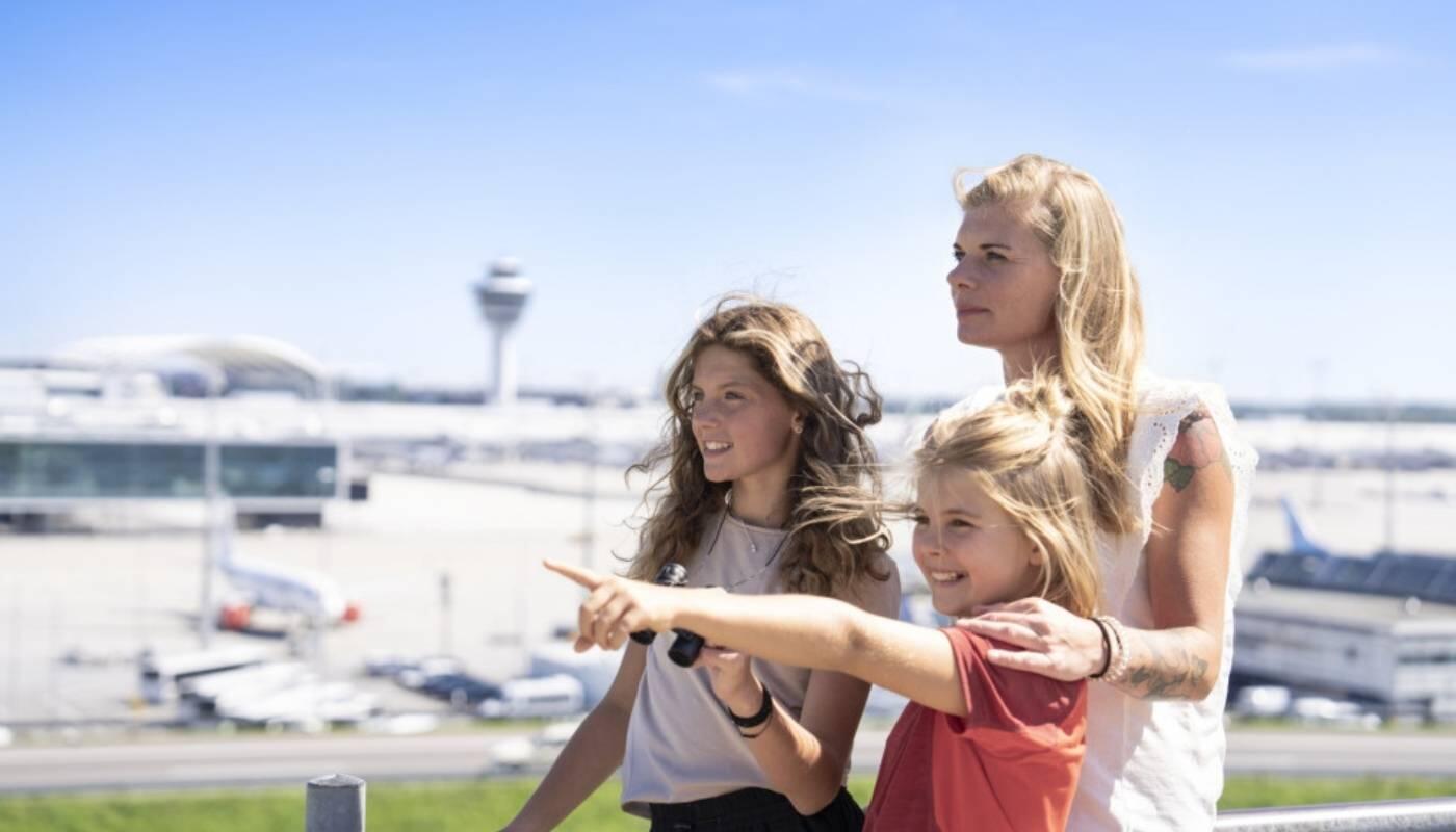 Family Time at the Visitor Park A mother with two daughters is standing on the visitors' hill, pointing into the distance. In the background, you can see the tarmac and the control tower against a bright blue sky.
