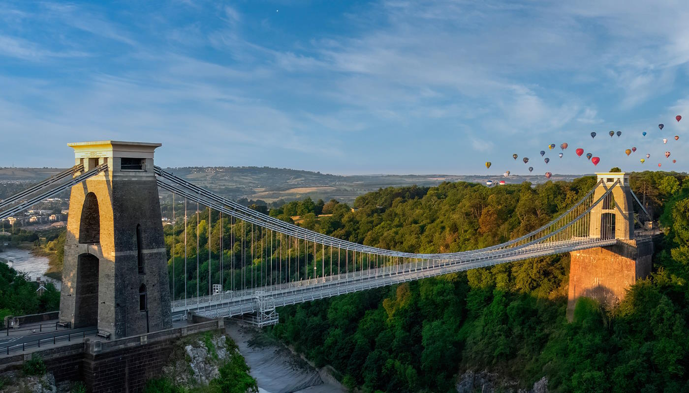 Clifton Brücke in Bristol, Großbritannien Das Bild zeigt die Clifton Suspension Bridge in Bristol, England, eine bekannte Hängebrücke über die Avon-Schlucht. Im Hintergrund sind bewaldete Hügel zu sehen, über denen zahlreiche bunte Heißluftballons am Himmel schweben. Die Brücke ist eine elegante Hängebrücke aus Stein und Stahl mit zwei massiven Pfeilern, zwischen denen die Fahrbahn an langen Seilen hängt. Sie spannt sich in einem sanften Bogen über die tiefe Schlucht und wirkt dadurch leicht und filigran.