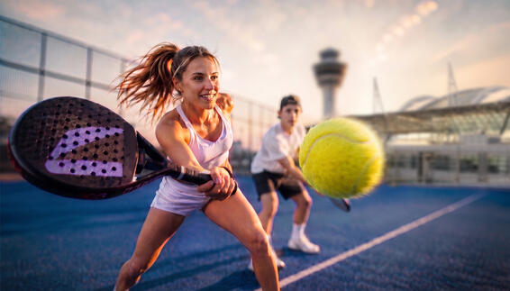 Dynamische Padel-Szene auf Outdoor-Court bei Sonnenuntergang mit Spieler beim kraftvollen Vorhand-Schlag, gelbem Ball im Vordergrund und weiterer Person im Hintergrund – am Flughafen München