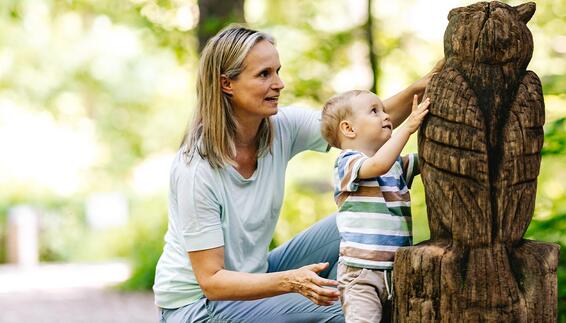 On the forest adventure trail in the Freising Forest, a woman is kneeling next to a toddler. The child reaches out and touches a large carved wooden owl.