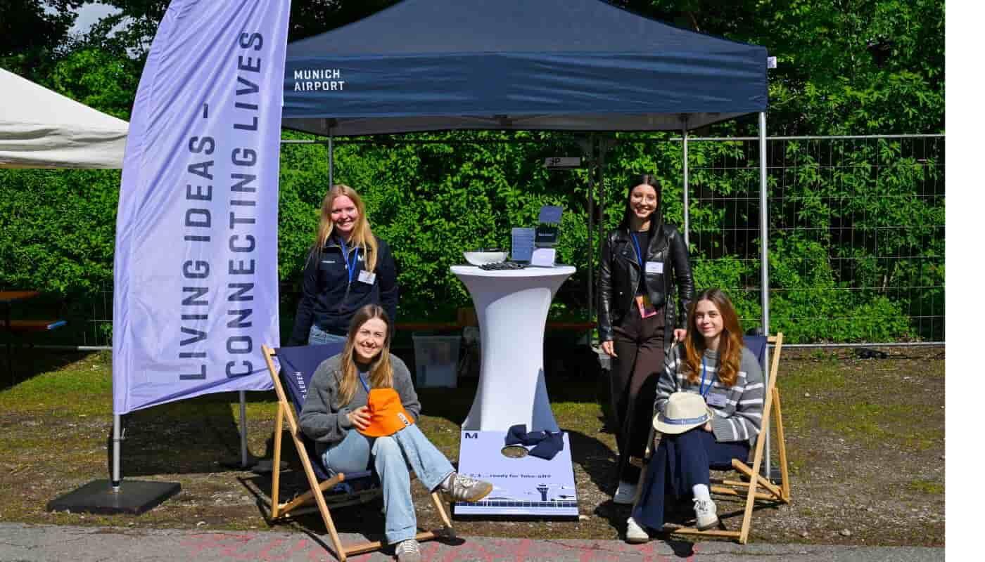 Munich Airport at the FünfSieben Festival – Supporting Young People and the Community Four young Munich Airport team members welcome visitors at the airport’s information booth during the FünfSieben Festival. In front of the pavilion displaying the message “Living Ideas – Connecting Lives,” the scene reflects a welcoming atmosphere of connection, community, and youth engagement.