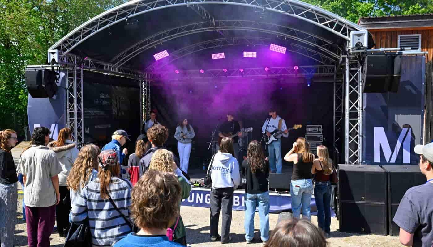 FünfSieben Festival at Karlsfelder See – Munich Airport Supports Youth Culture Young festivalgoers enjoy the vibrant atmosphere of the FünfSieben Festival in front of an outdoor stage at Karlsfelder See, where a live band performs, bringing together music, community, and youth culture.