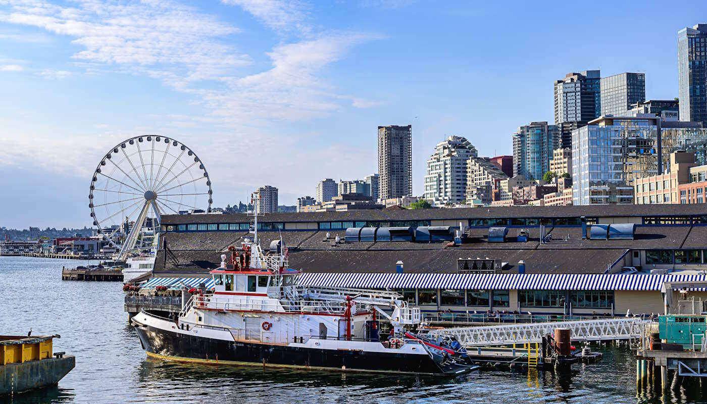 Waterfront in Seattle, USA Das Bild zeigt die Waterfront von Seattle mit einem großen Riesenrad, einer Anlegestelle und einem Schiff im Vordergrund. Dahinter erhebt sich die moderne Skyline der Stadt entlang des Puget Sound.
