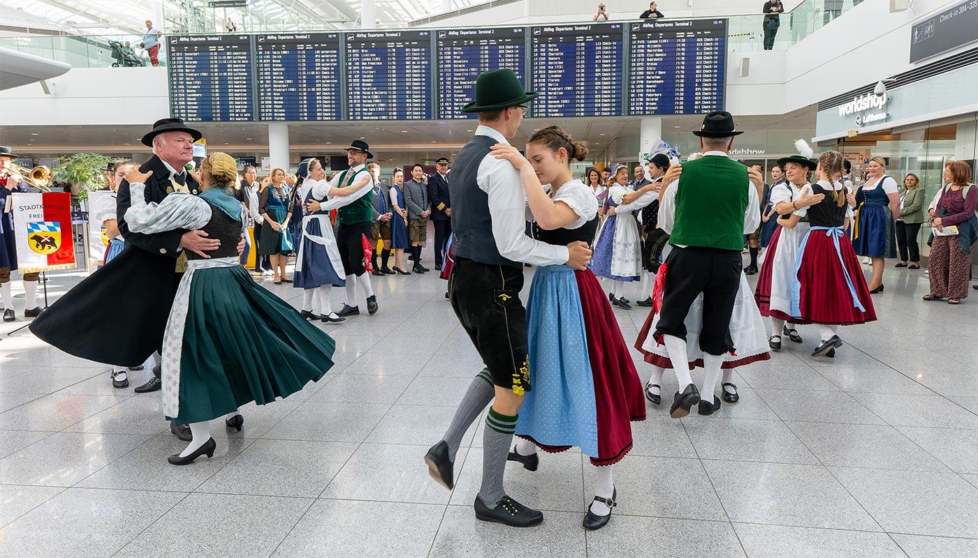 Oktoberfest 2026 Group of people in traditional Bavarian dress dancing in an airport terminal, with a large departure board and waiting travelers in the background.