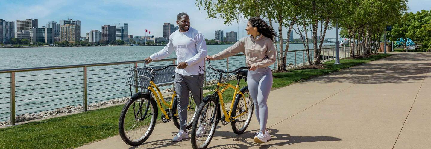 Detroit Zwei Personen schieben Fahrräder auf einem Fußweg entlang des Detroit River mit Blick auf die Skyline von Detroit und das Wasser im Hintergrund.